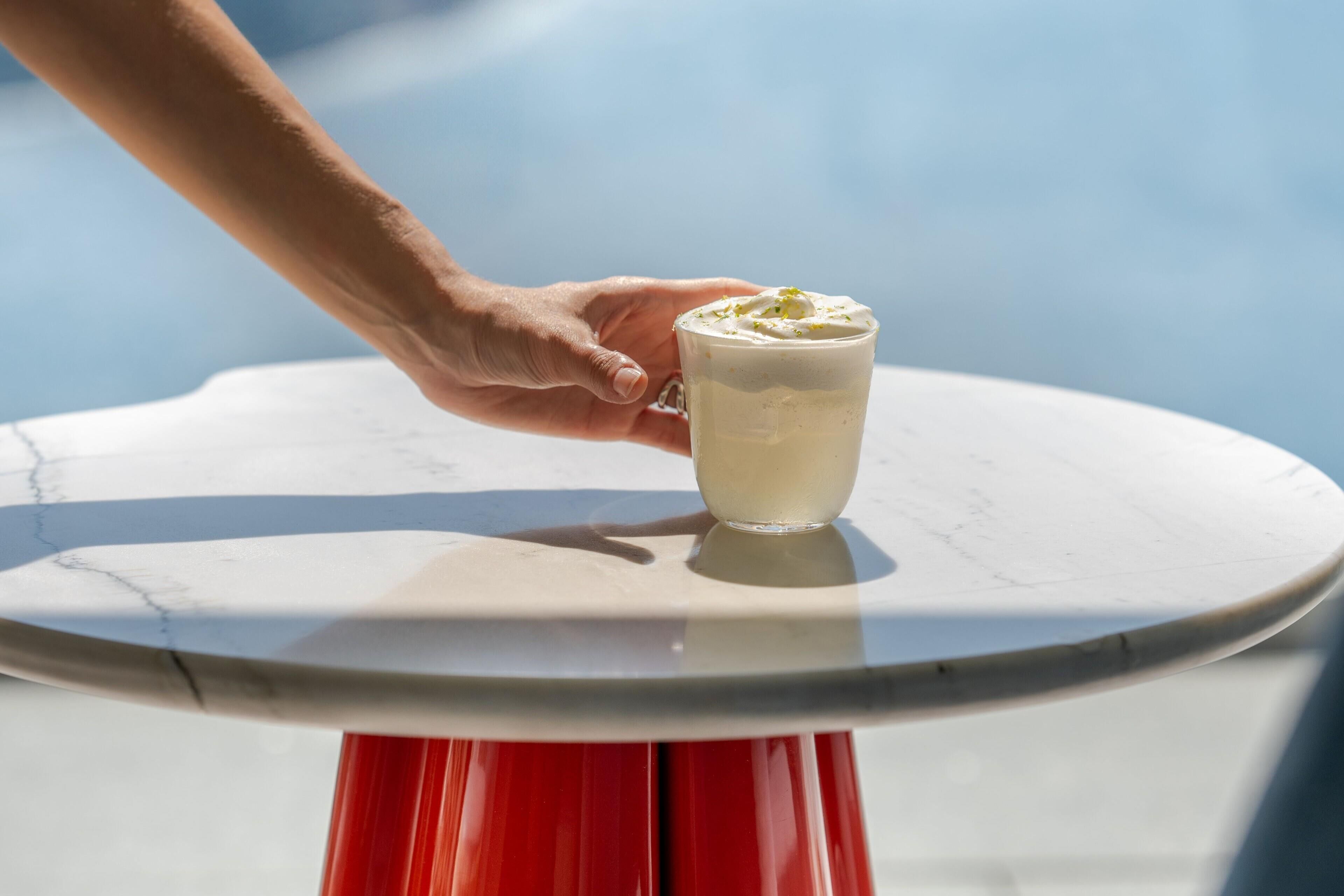 A hand holding a glass of white beverage on a white table with a red base.