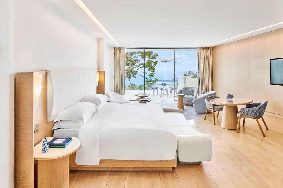 A modern hotel room with a large bed, a seating area with a table, and a view of the sea.