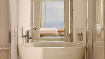 Un baño moderno con una bañera independiente grande, un box de ducha y vistas a un dormitorio con cama y ventana con vista al mar.