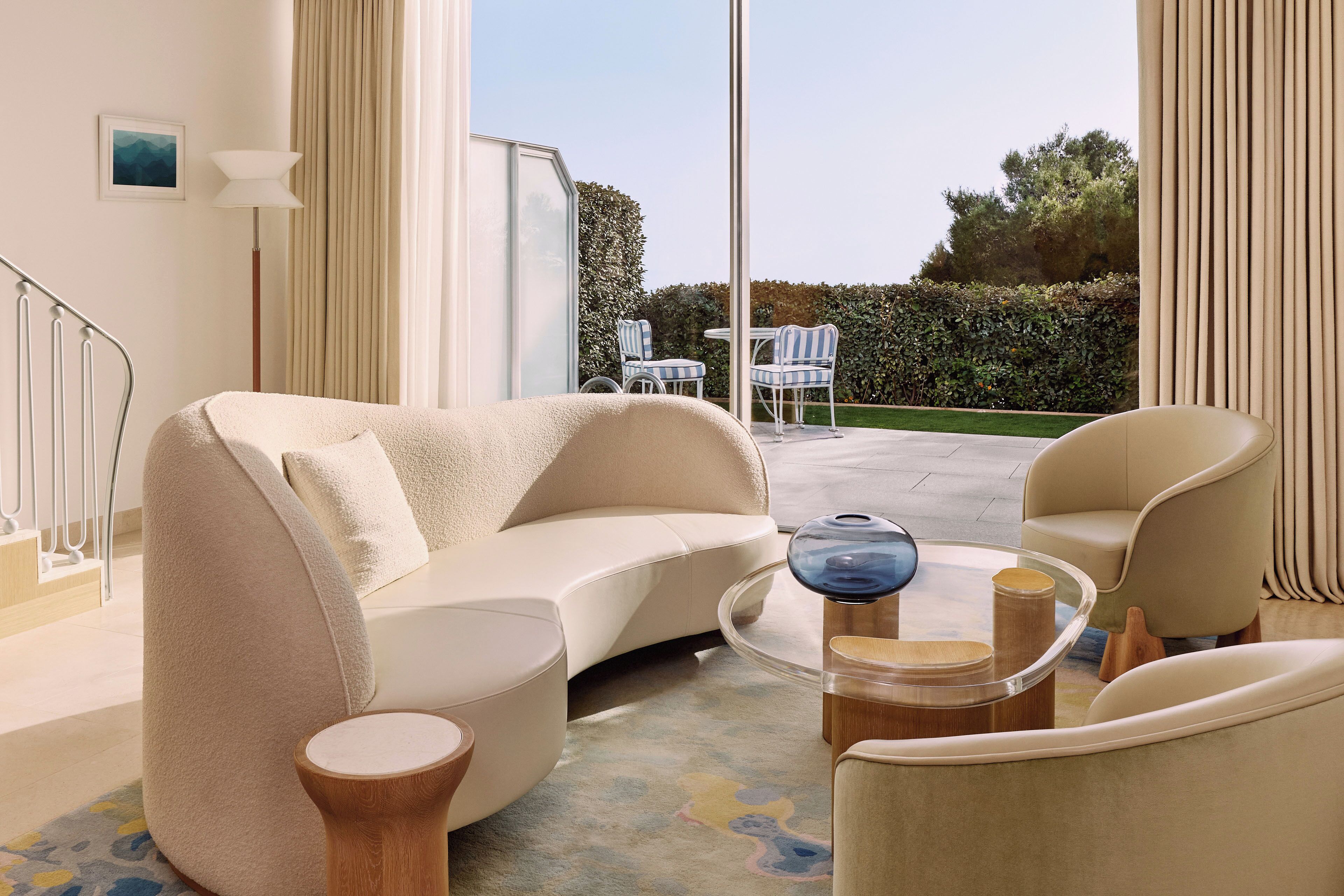 A modern living room with a curved sofa, a round coffee table, and two armchairs. A staircase is visible in the background leading to an outdoor area with a table and chairs.