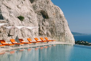 A pool with lounge chairs and umbrellas, a rocky cliff, and a view of the sea. - The Maybourne Riviera, Maybourne  (Roquebrune-Cap-Martin)
