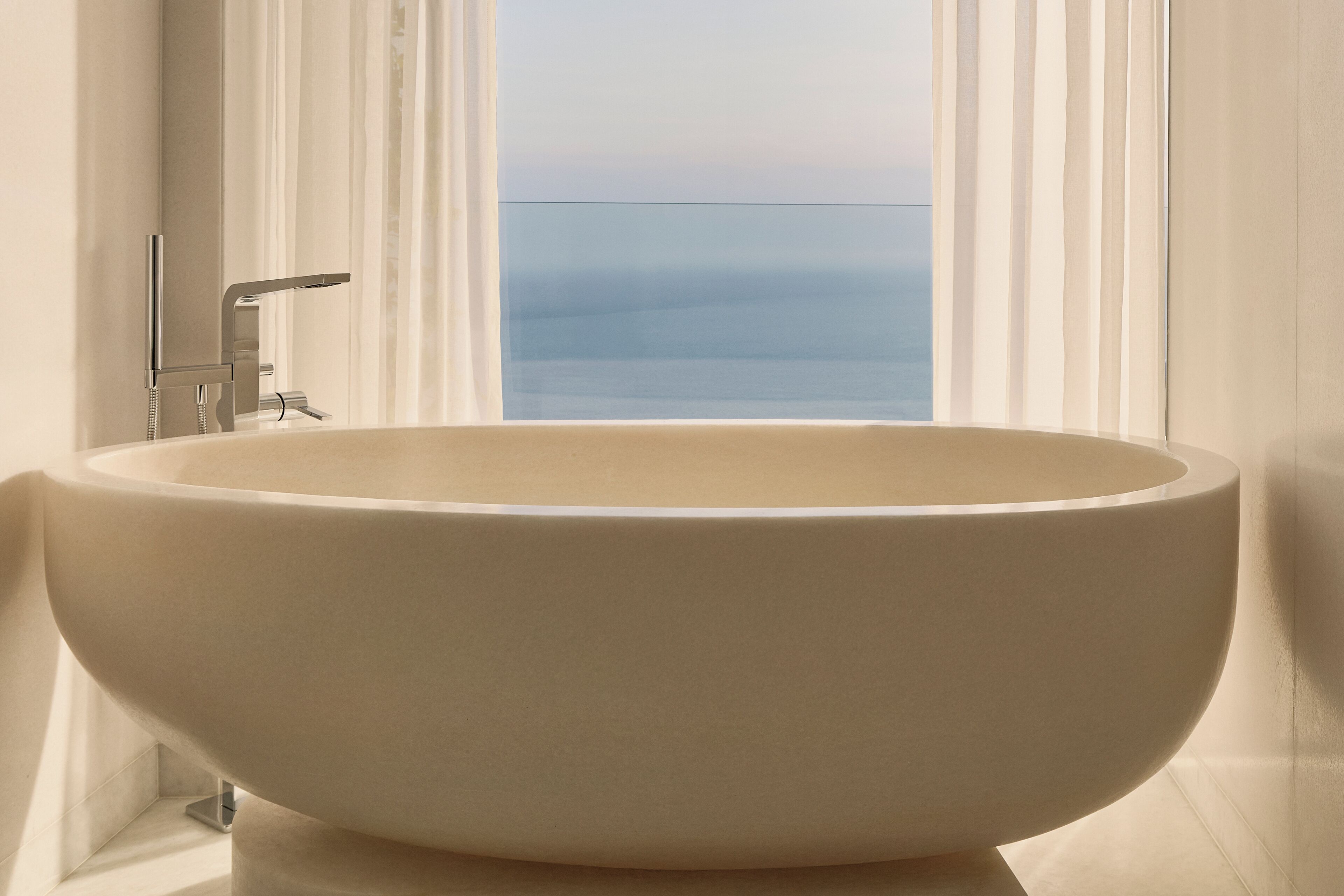 A large, oval-shaped bathtub in a bathroom with a view of the sea through a window.