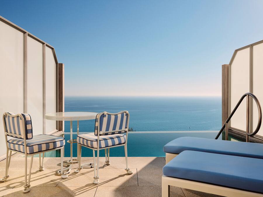 Un balcone con due sedie a righe, un tavolino e una panca imbottita blu, con vista su un mare azzurro e limpido.