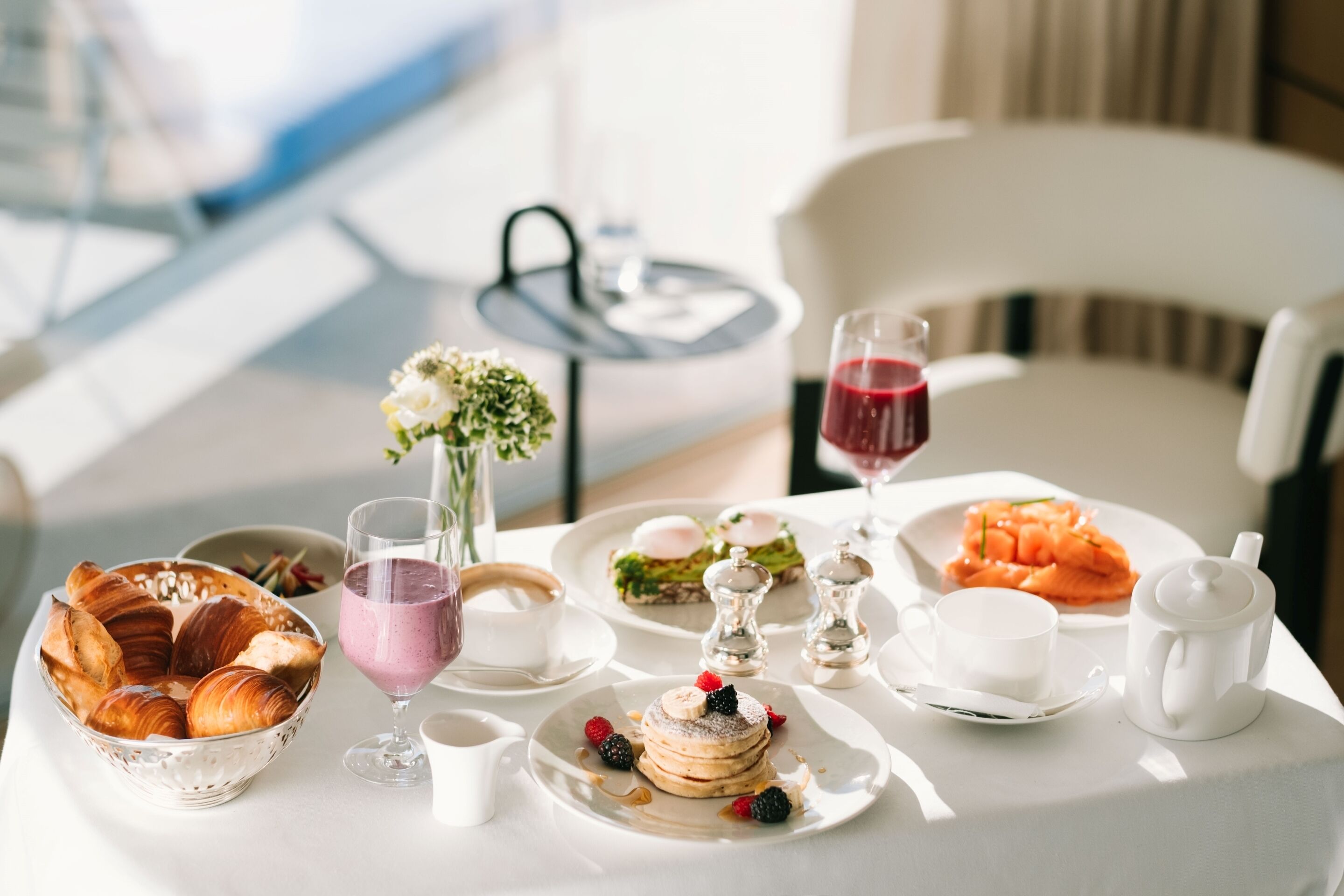 Un desayuno con cruasanes, un batido, tortitas con frutos rojos, salmón y una tetera sobre una mesa.