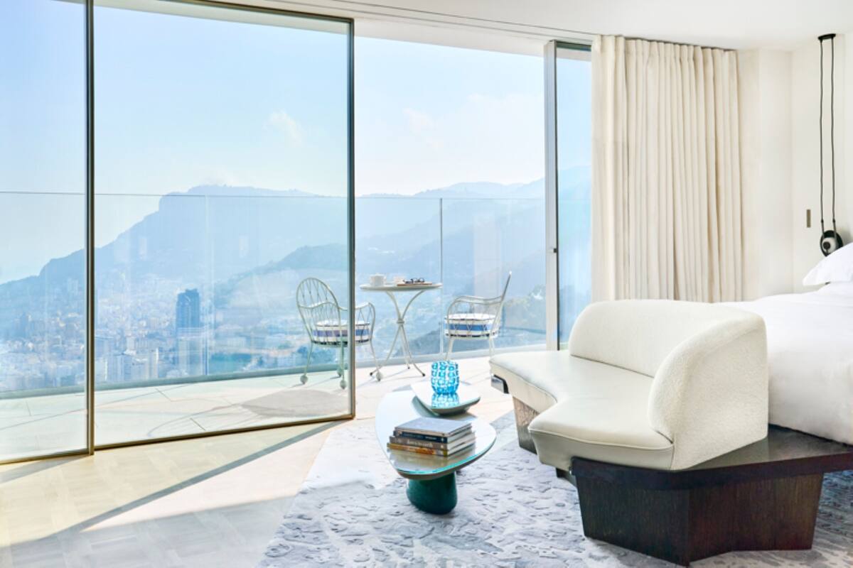 A modern hotel room with a large sliding glass door, a white sofa, a small round table with books, and a view of a cityscape.