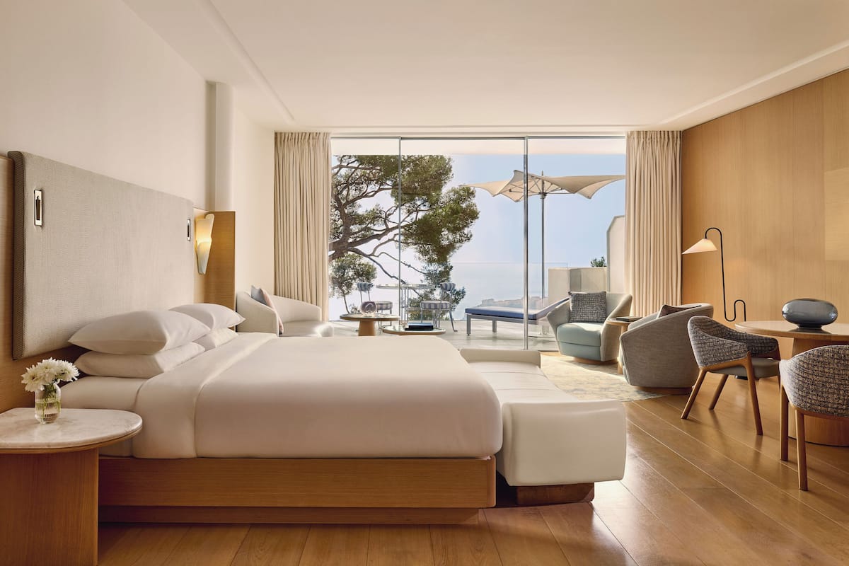 A modern hotel room with a large bed, a sitting area with a view of the sea, and a balcony with outdoor furniture.
