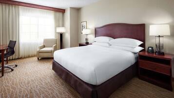 Habitación de hotel con una cama grande, un escritorio con silla, una mesita de noche con lámpara y una ventana con cortinas.