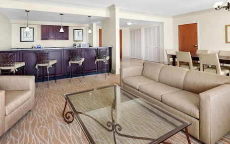 A modern living room with a beige sofa, a glass coffee table, and a kitchen area with a bar and bar stools.