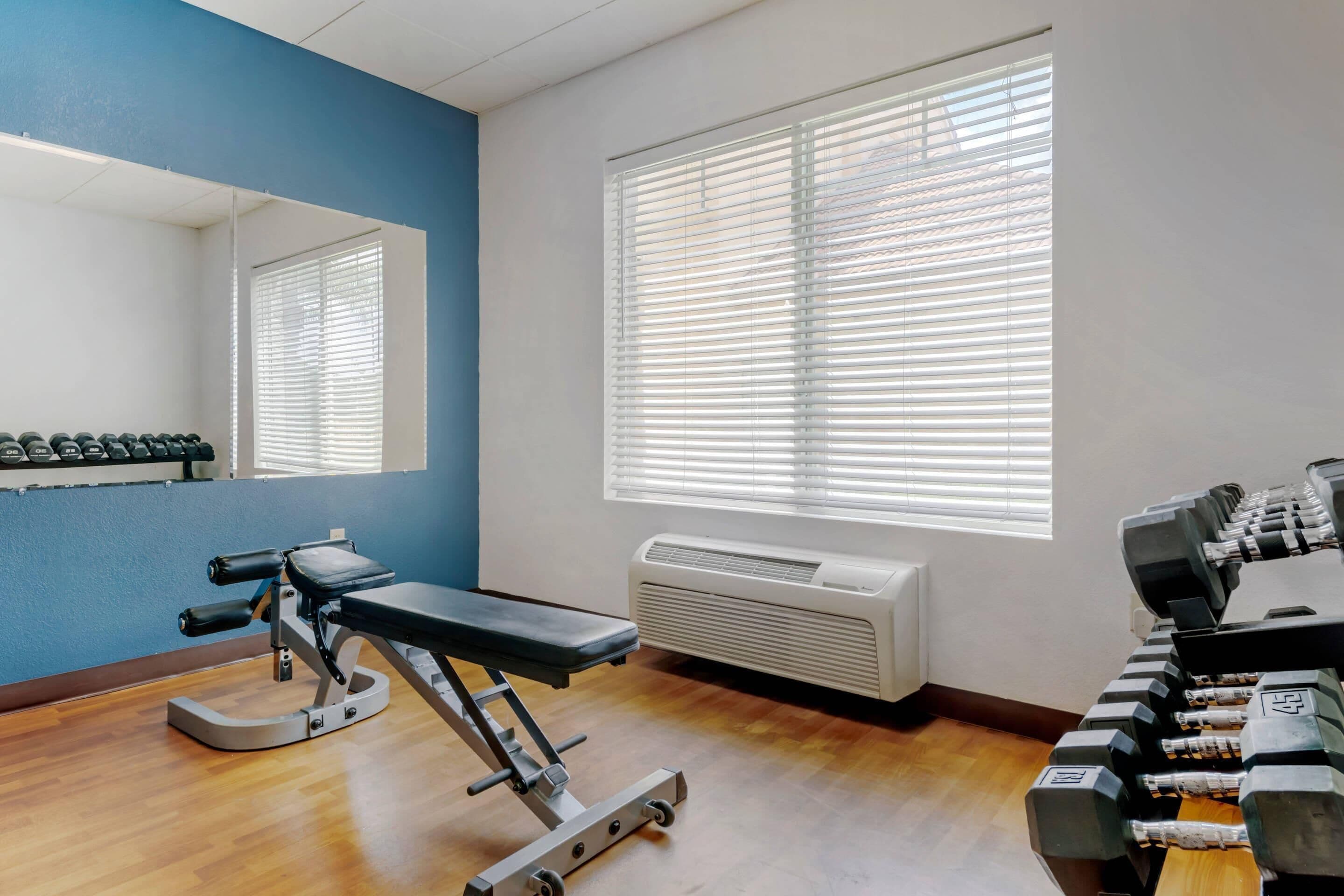 Een sportschool met trainingsapparatuur, een spiegel en grote ramen met zonwering.