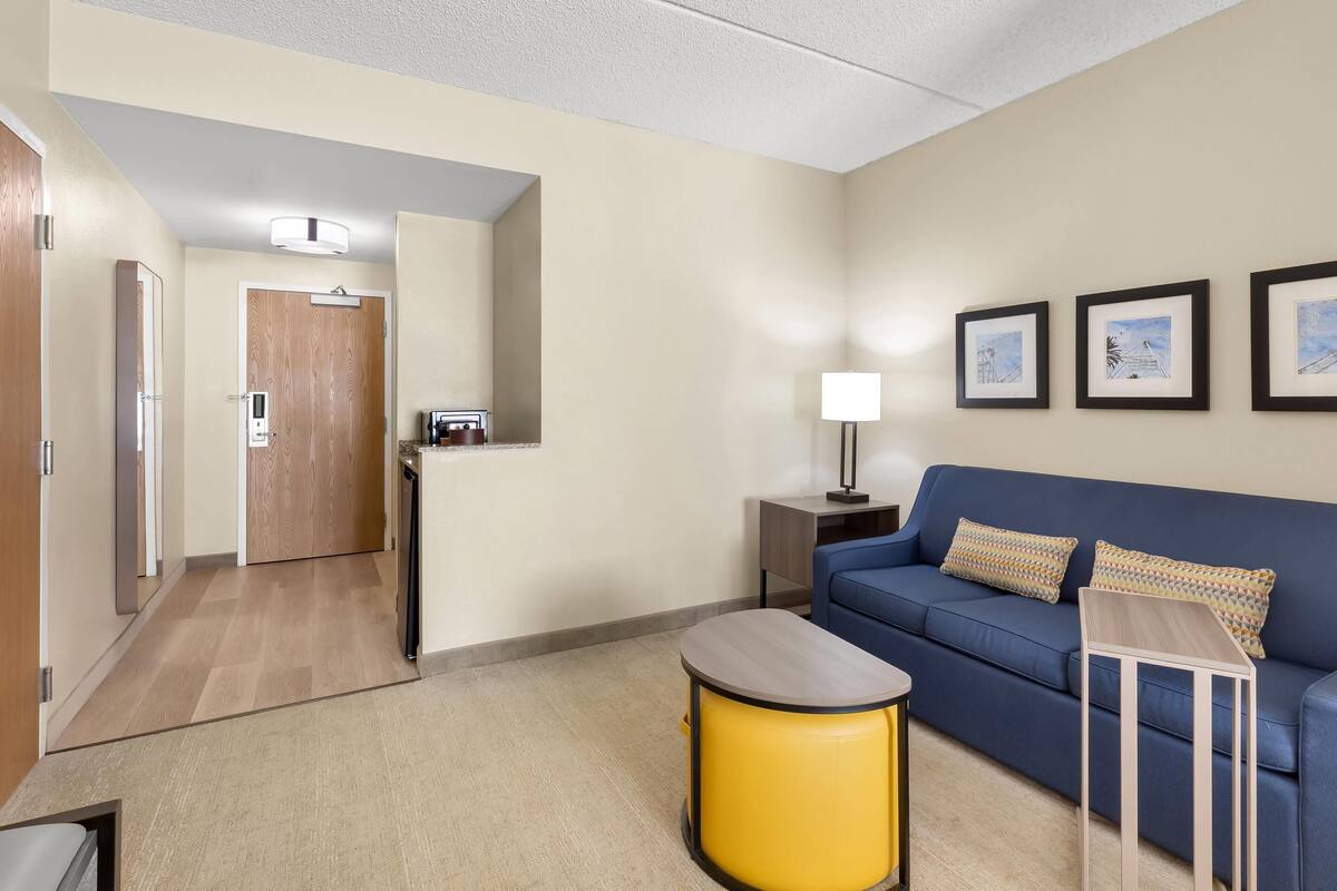 Une chambre d’hôtel moderne avec un canapé bleu, une petite table en bois, un pouf jaune et des tableaux encadrés accrochés au mur.