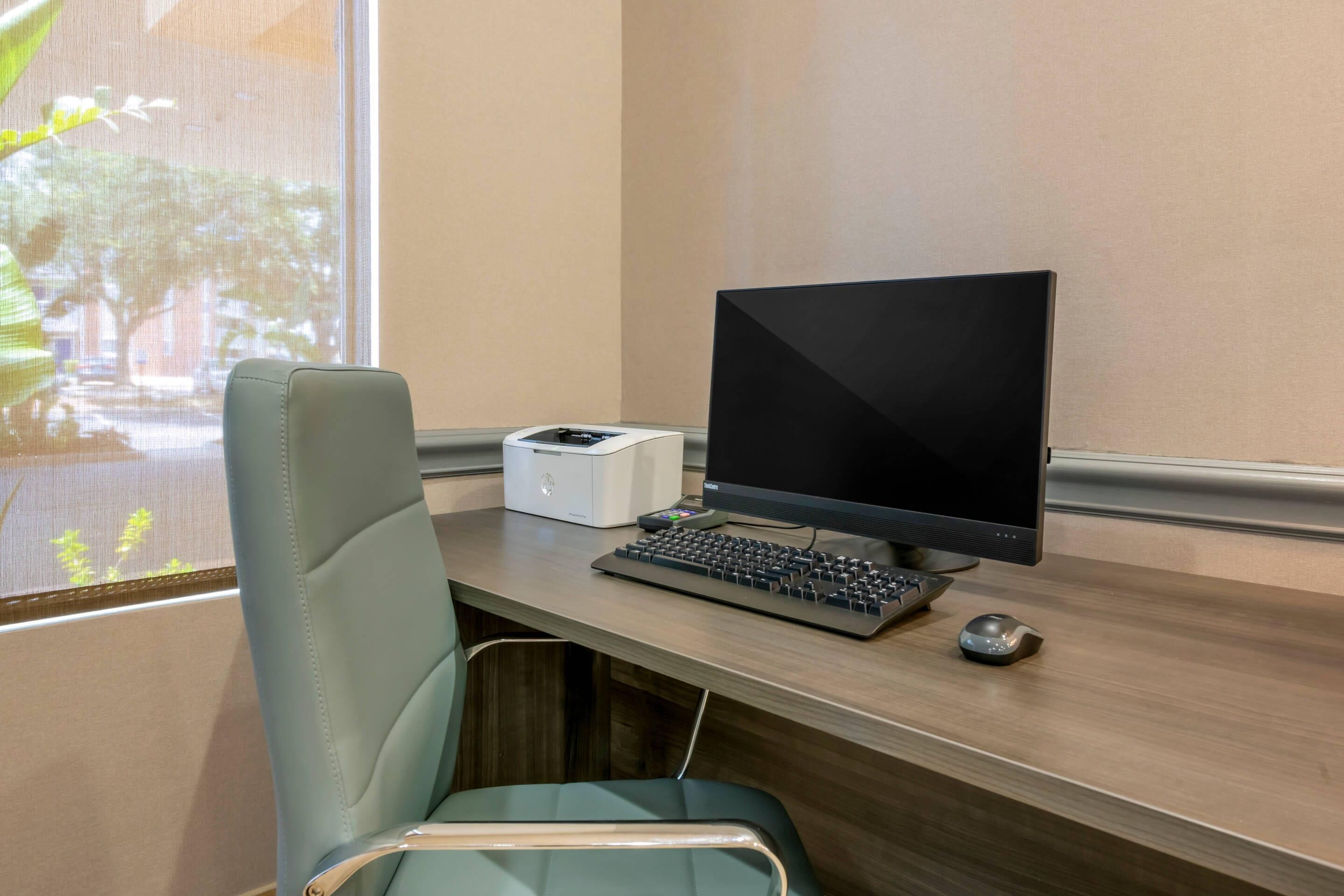 A modern office chair, a desktop computer with a monitor, keyboard, and mouse, and a printer on a wooden desk.
