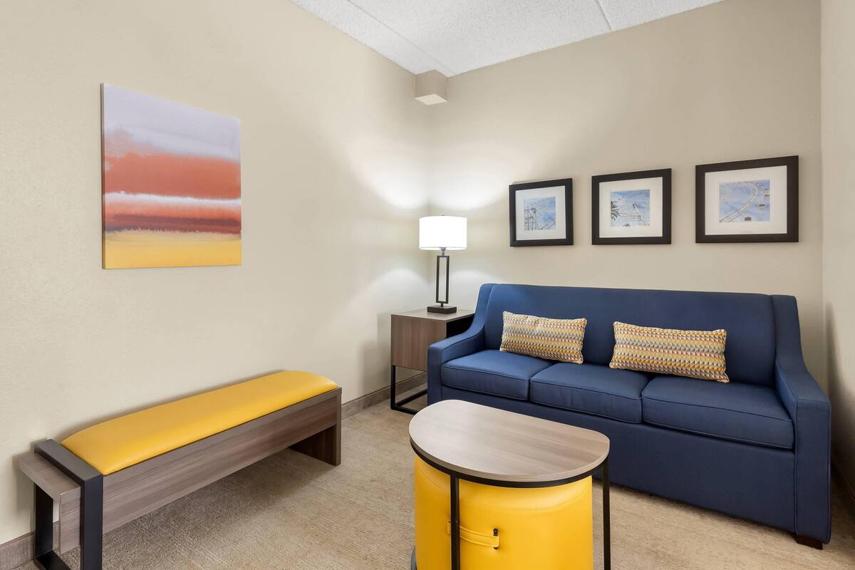 Un salon moderne avec un canapé bleu, un banc jaune et une table ronde. Trois tableaux encadrés ornent le mur, et une lampe sur pied est placée dans un coin.
