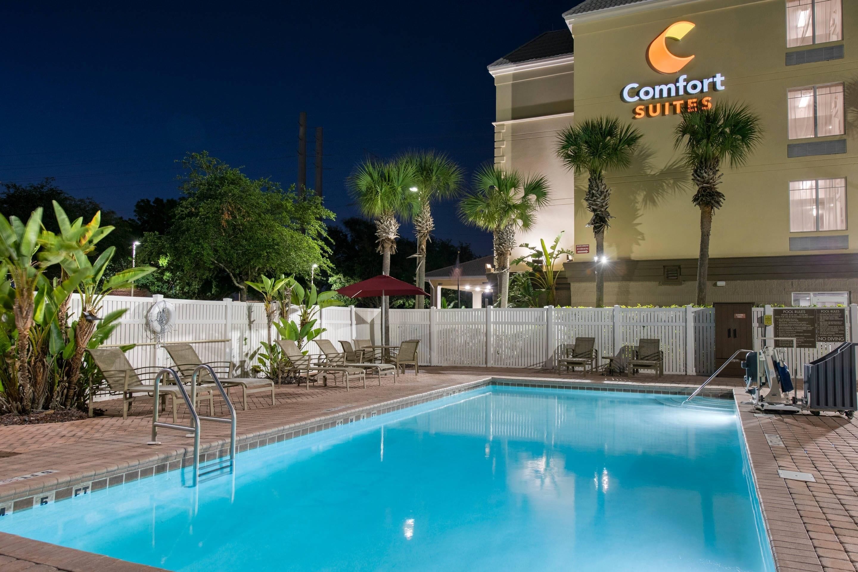 A hotel pool area with lounge chairs, palm trees, and a Comfort Suites building in the background.