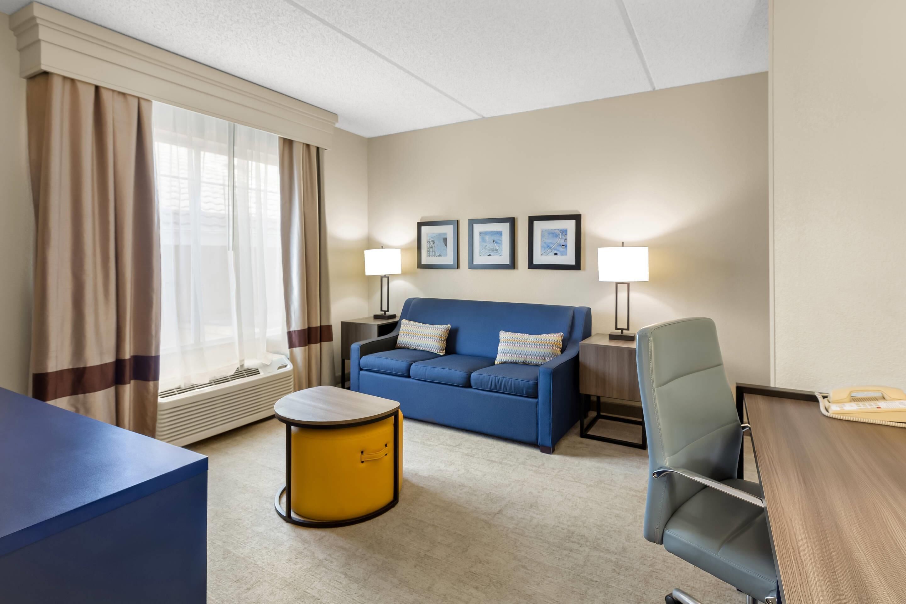Une chambre d’hôtel avec un canapé bleu, une table d’appoint jaune, une chaise bleue, un bureau en bois et une fenêtre avec des rideaux.