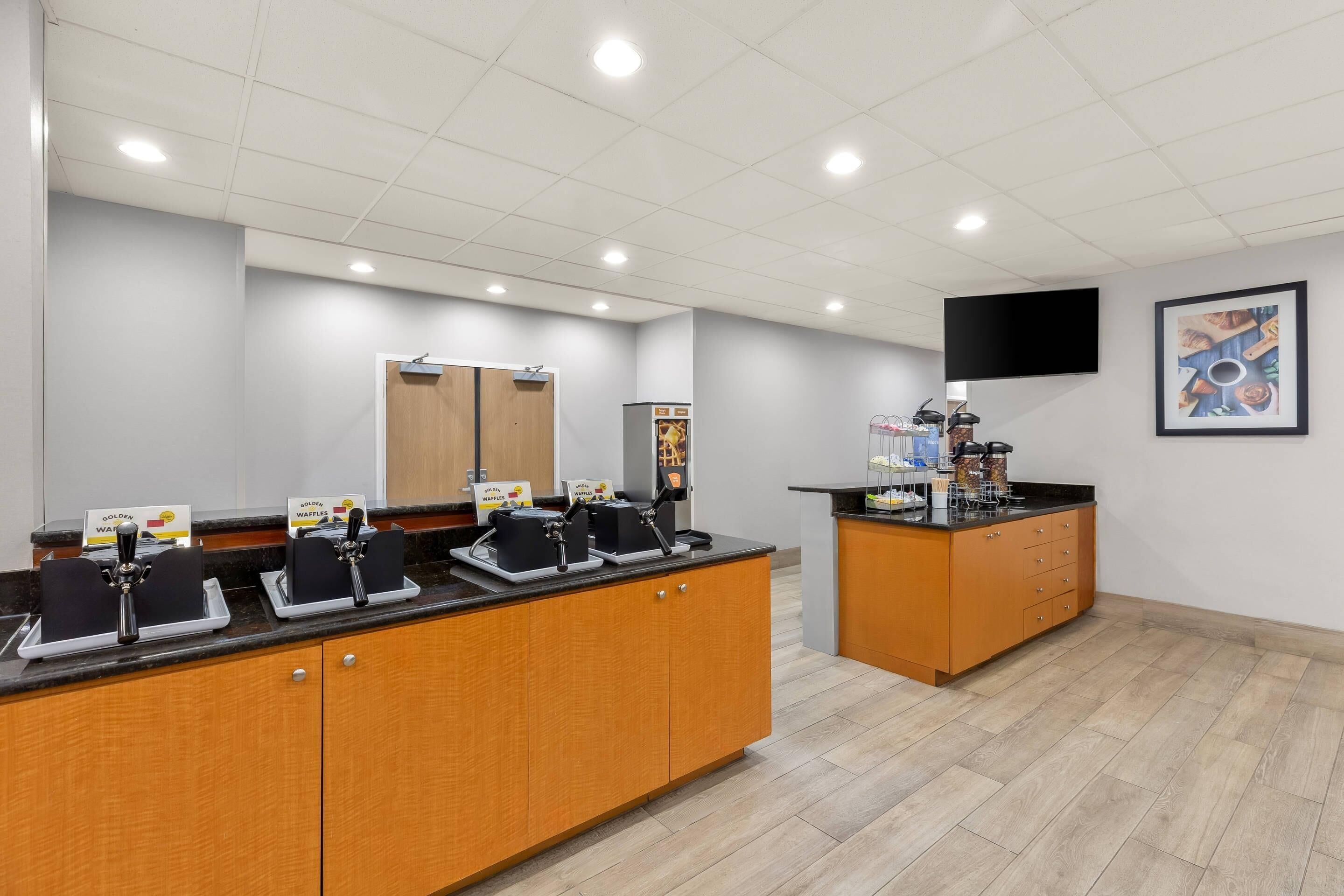 A modern kitchen with a wooden counter, two waffle stations, a coffee machine, and a flat-screen TV on the wall.