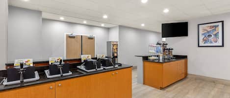 A modern kitchen with a wooden counter, two waffle stations, a coffee machine, and a flat-screen TV on the wall.