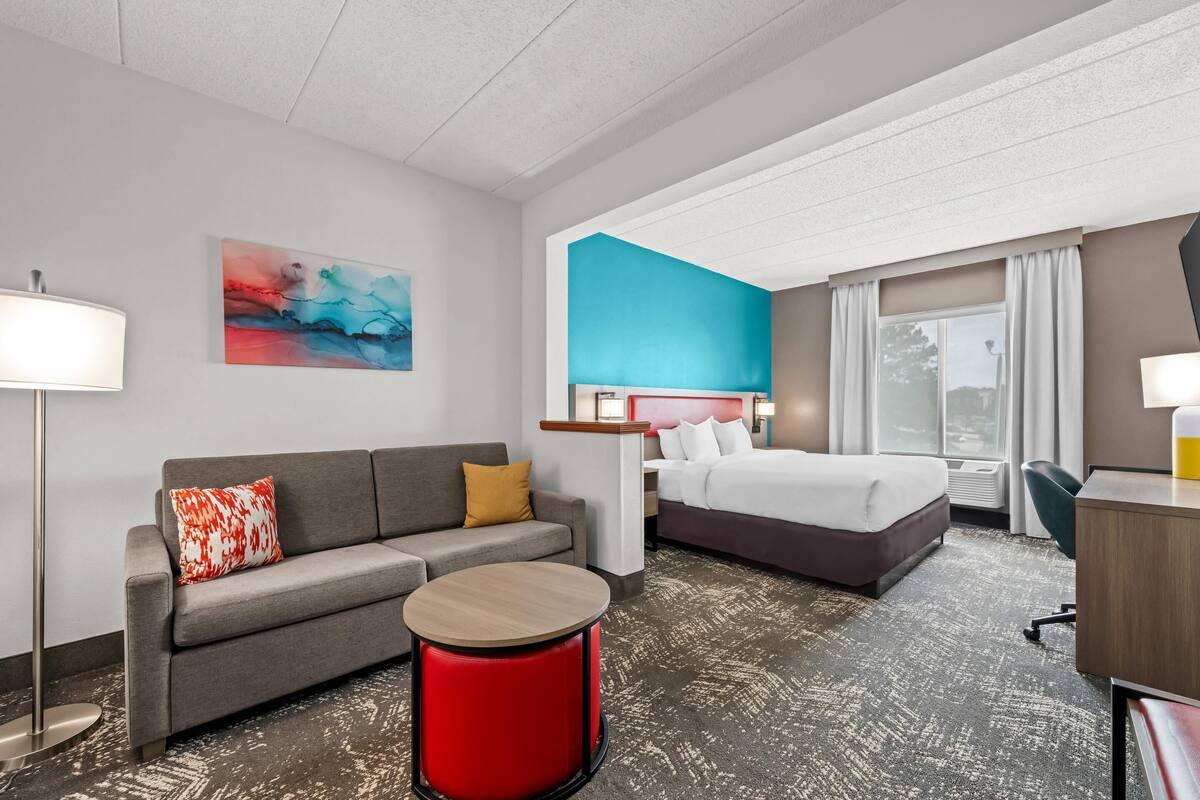 A hotel room with a grey sofa, a red accent stool, a bed with white bedding, a desk, and a large window with curtains.