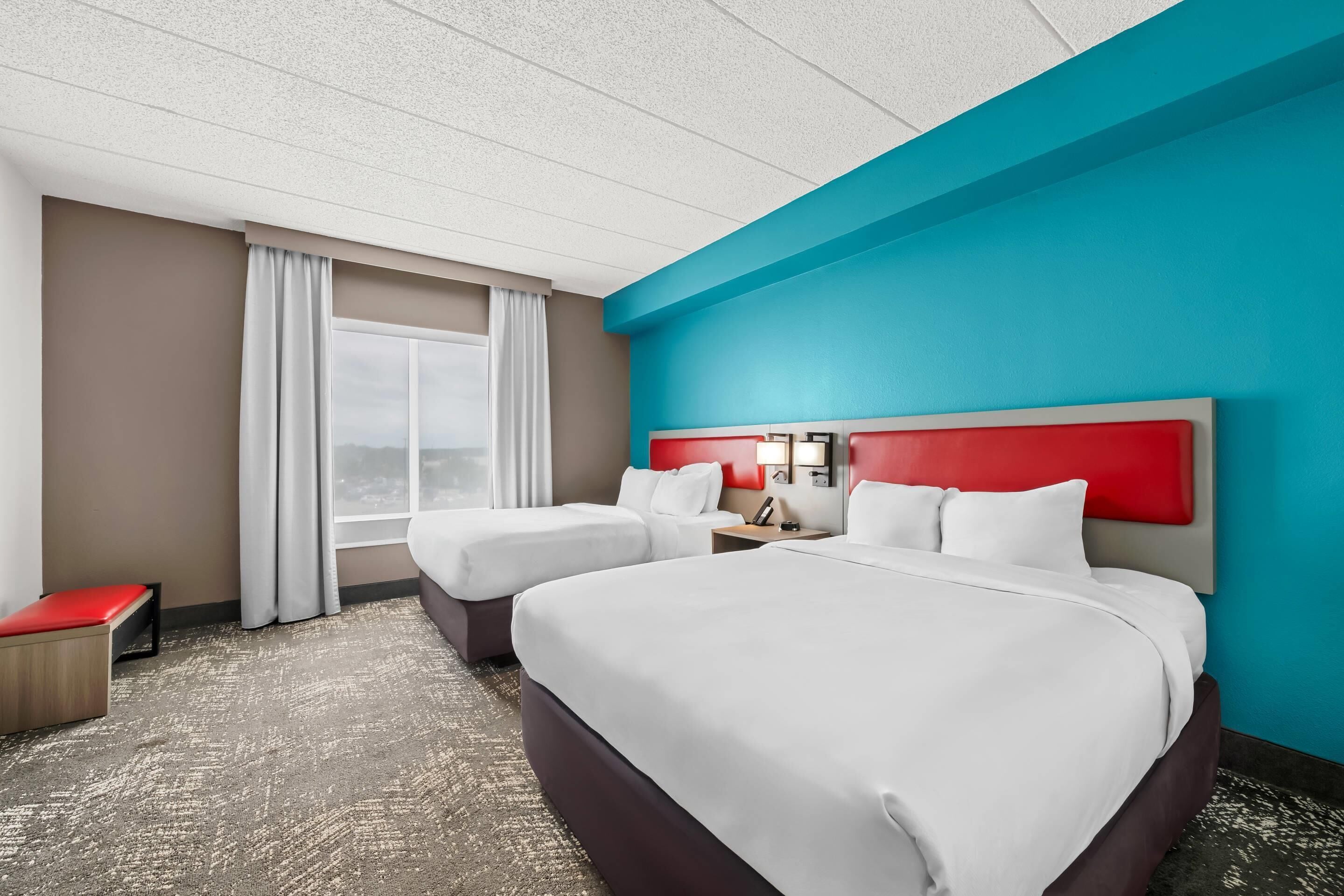 A hotel room with two beds, a red bench, a window with curtains, and a view of the outdoors.