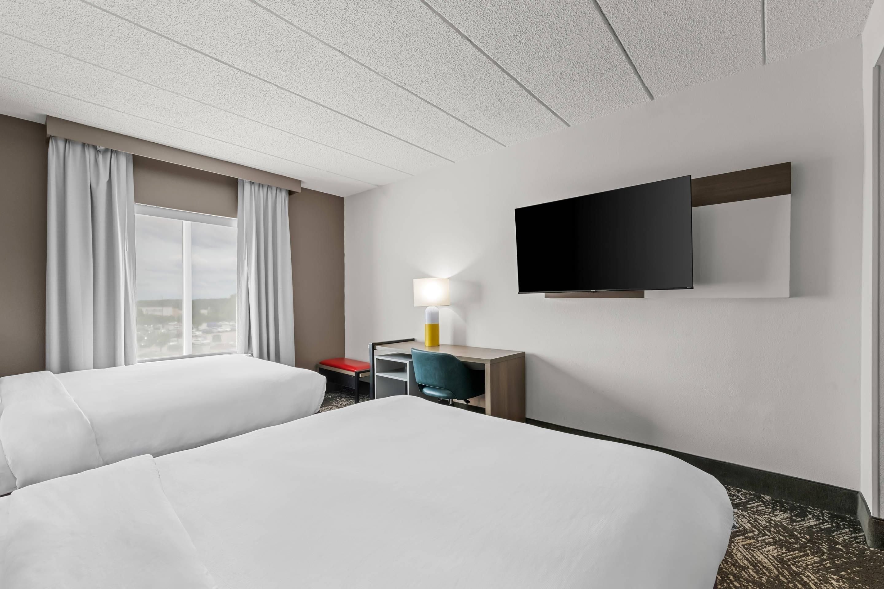 A hotel room with two beds, a desk, a chair, a TV, and a window with curtains.