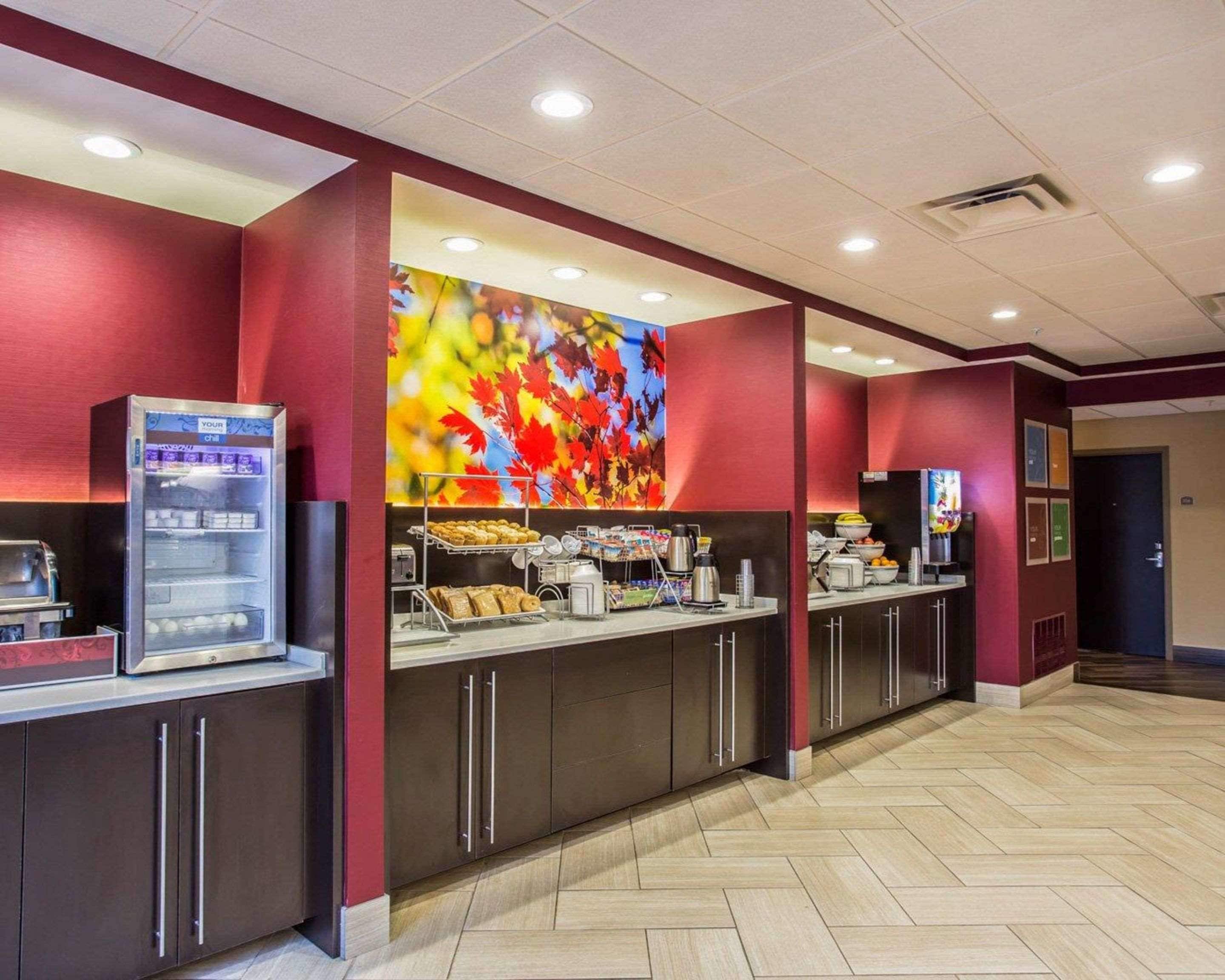 A hotel lobby with a self-service breakfast area, including a vending machine, a coffee station, and a large colorful mural on the wall.