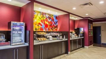 A hotel lobby with a self-service breakfast area, including a vending machine, a coffee station, and a large colorful mural on the wall.