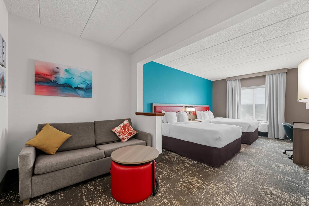 A hotel room with a grey sofa, a red ottoman, a bed with white bedding, a blue accent wall, and a painting on the wall.
