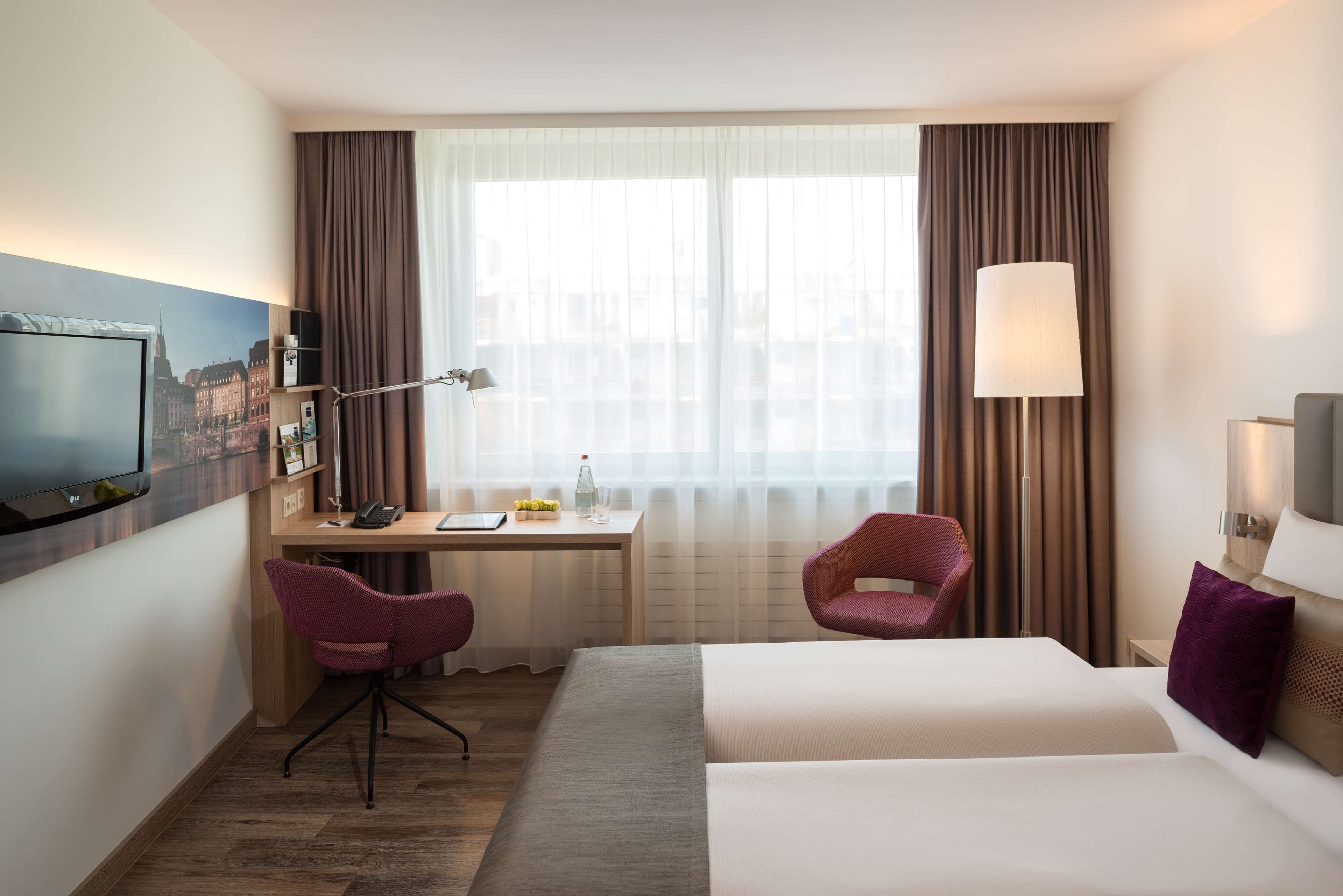 A hotel room with a bed, a desk with a chair, a TV, and a window with curtains.
