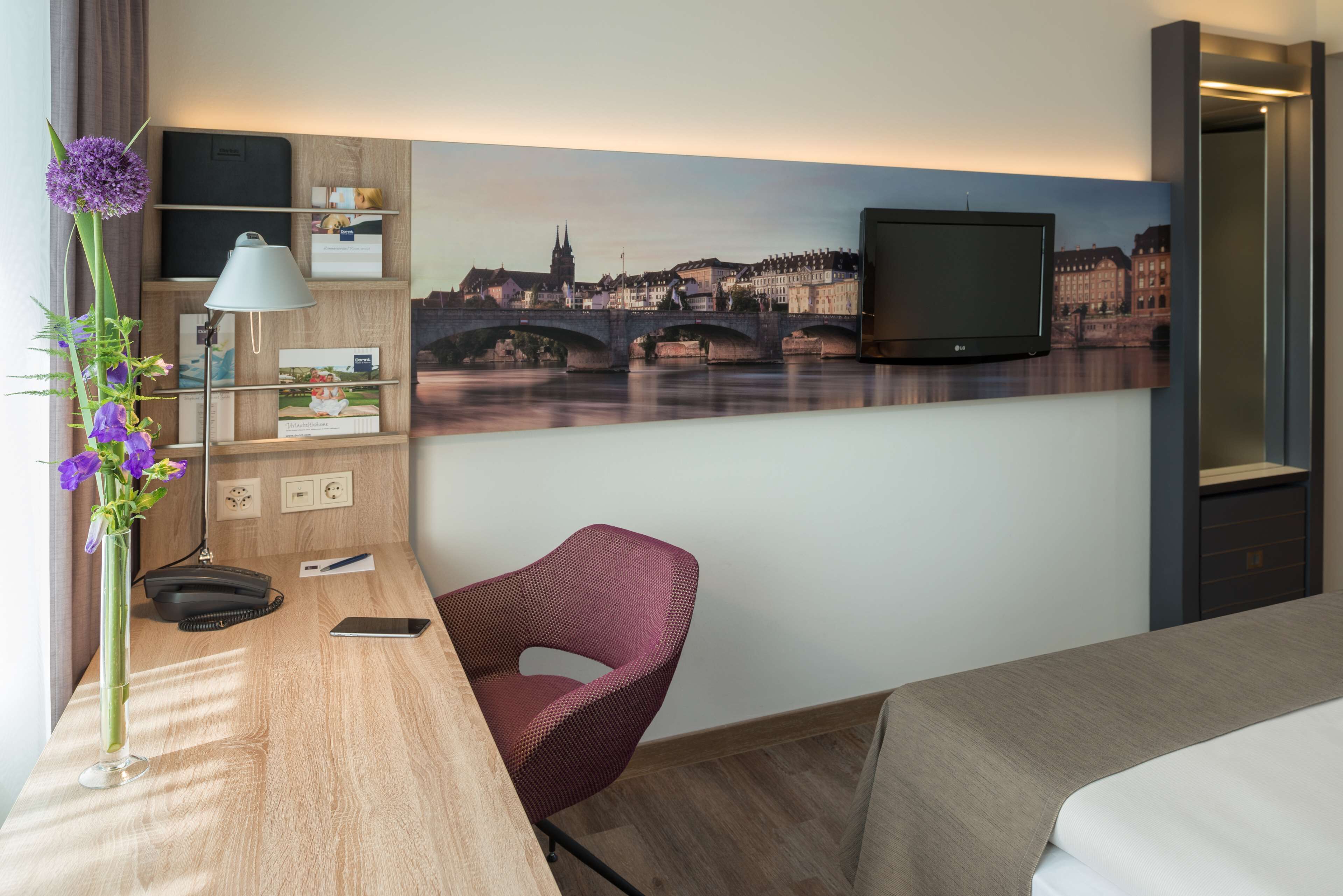Una camera d'albergo con un letto, una scrivania con una lampada, una sedia, un televisore montato a parete e un quadro con una veduta sulla città.