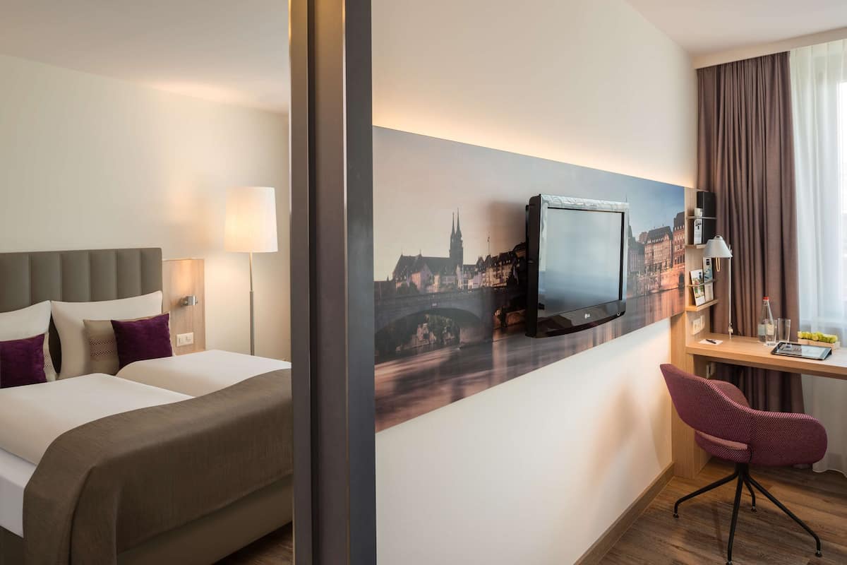 A hotel room with a bed, a desk, a chair, a television, and a cityscape painting on the wall.