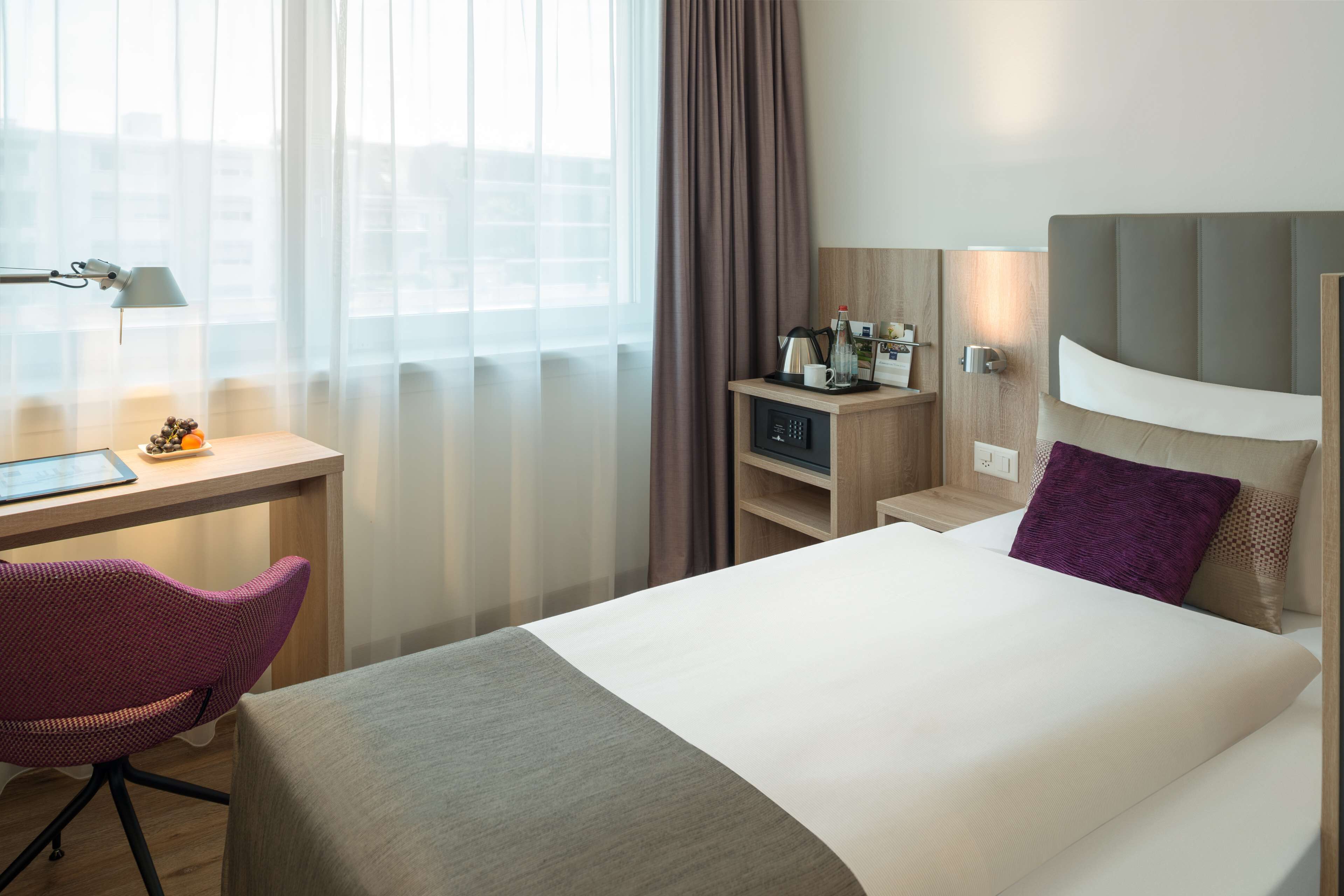 Habitació d'hotel amb llit, escriptori i cadira, una làmpada i finestra amb cortines.