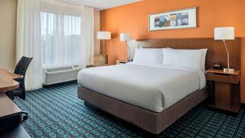 Quarto de hotel com uma cama grande, duas mesas de cabeceira com luminárias, uma cadeira, uma escrivaninha e uma janela com cortinas.