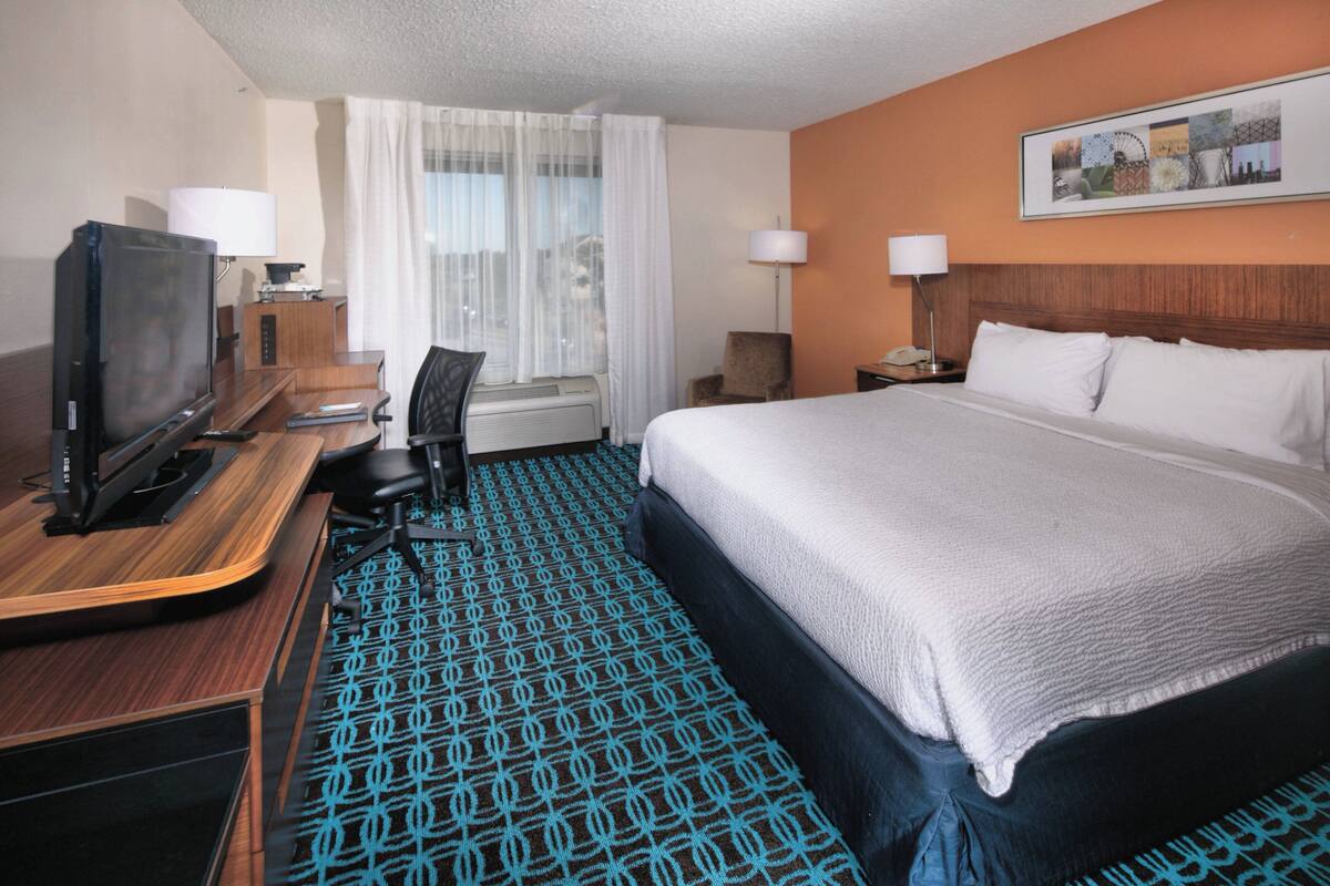 A hotel room with a large bed, a desk with a television, a chair, and a desk lamp.