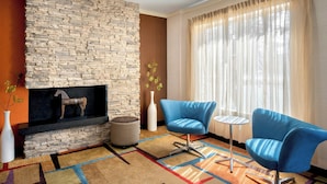 A modern living room with a stone fireplace, blue chairs, a patterned rug, and a large window with sheer curtains. - Fairfield Inn & Suites by Marriott Dover (Dover)