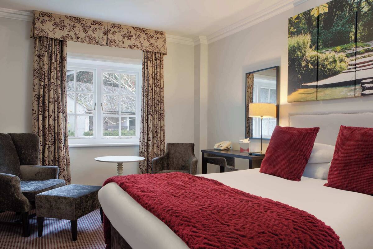 A hotel room with a bed, two armchairs, a small table, a lamp, a phone, and a painting on the wall.