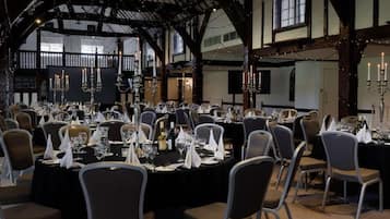 Un salón de banquetes con mesas redondas dispuestas para cenar, con candelabros y un techo alto con vigas a la vista.