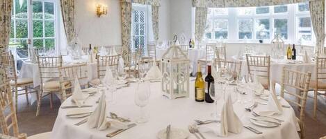 Comedor formal con mesas redondas dispuestas para una comida, manteles blancos y sillas de mimbre.