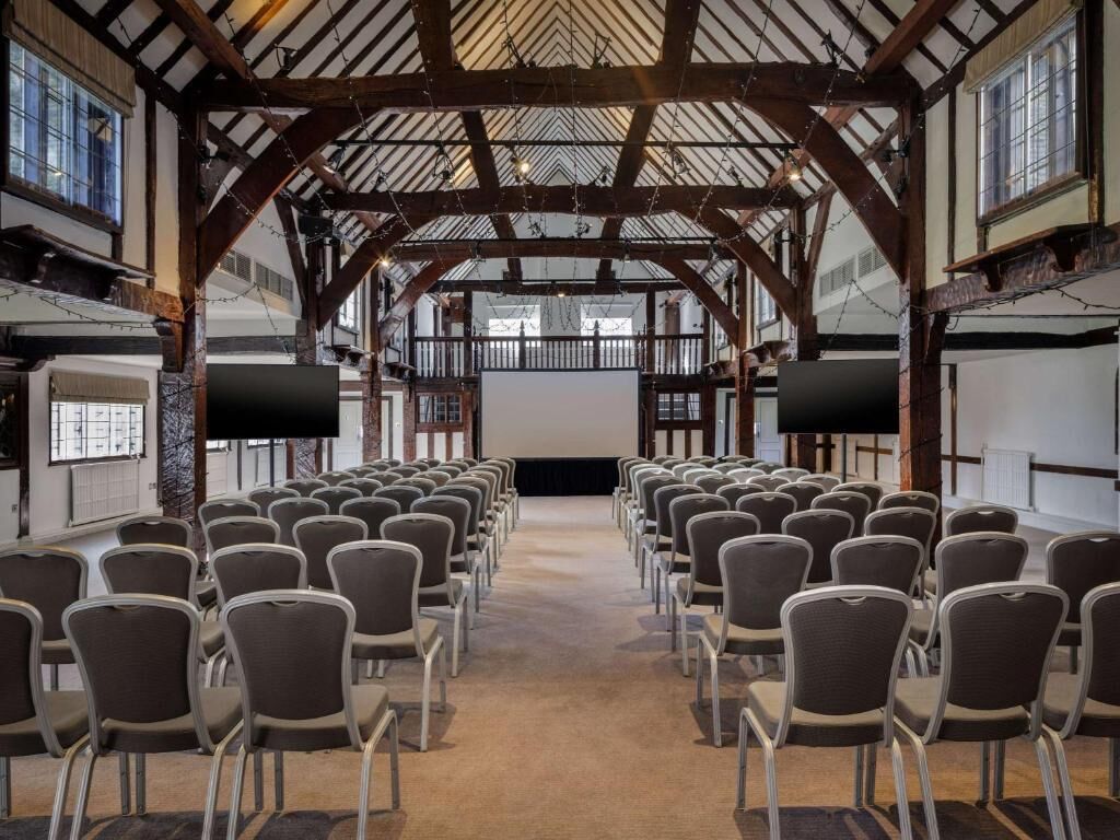 Une salle de conférence avec des rangées de chaises disposées face à un grand écran, des poutres en bois au plafond et des fenêtres le long des murs.