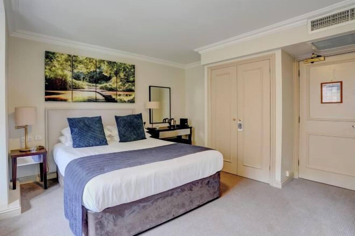 A hotel room with a bed, a nightstand, a lamp, a painting, and a door.