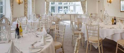 A banquet hall with round tables, white tablecloths, and wooden chairs.