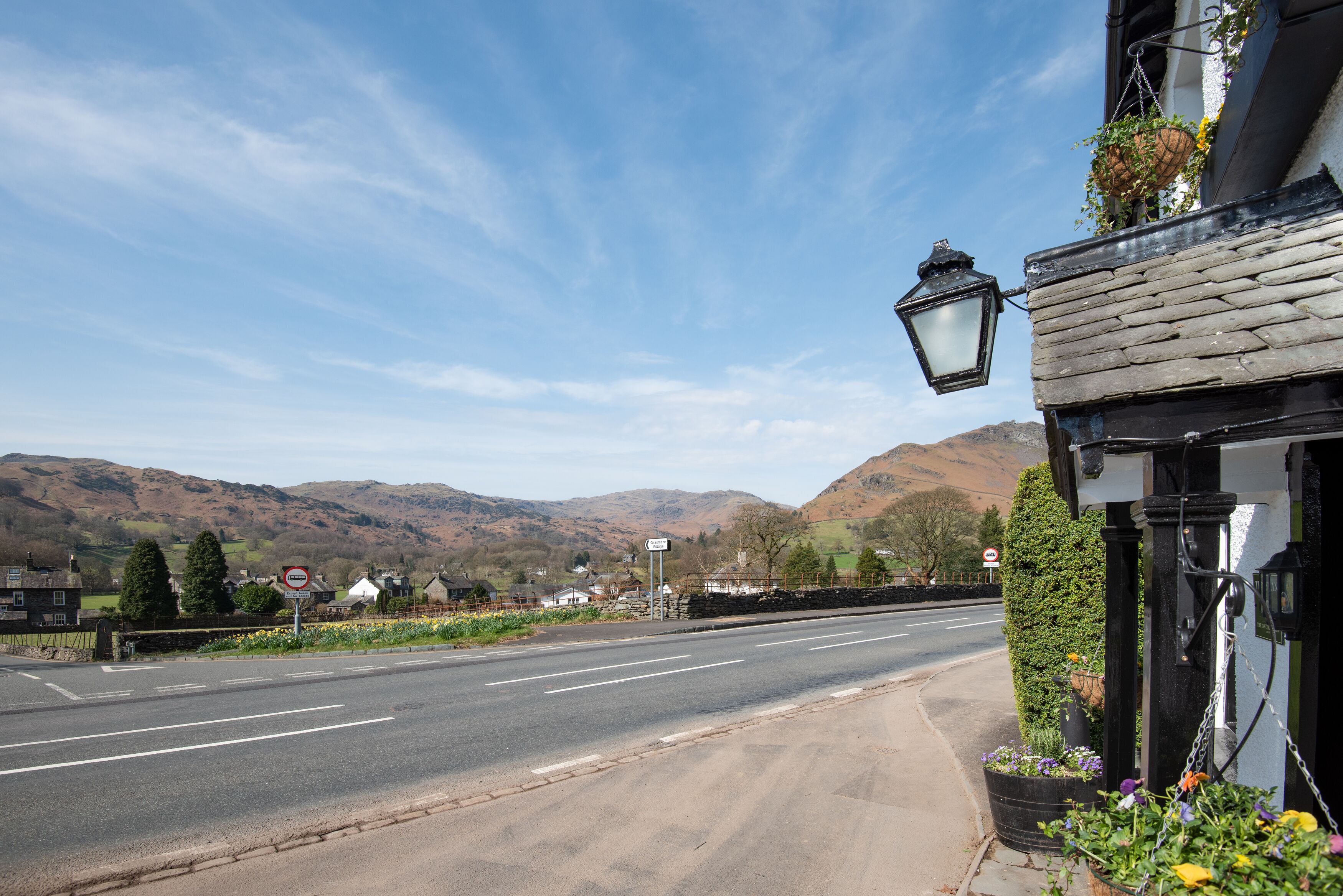 A road with a 'no entry' sign, a street lamp, and a building with a stone facade and hanging flower baskets.