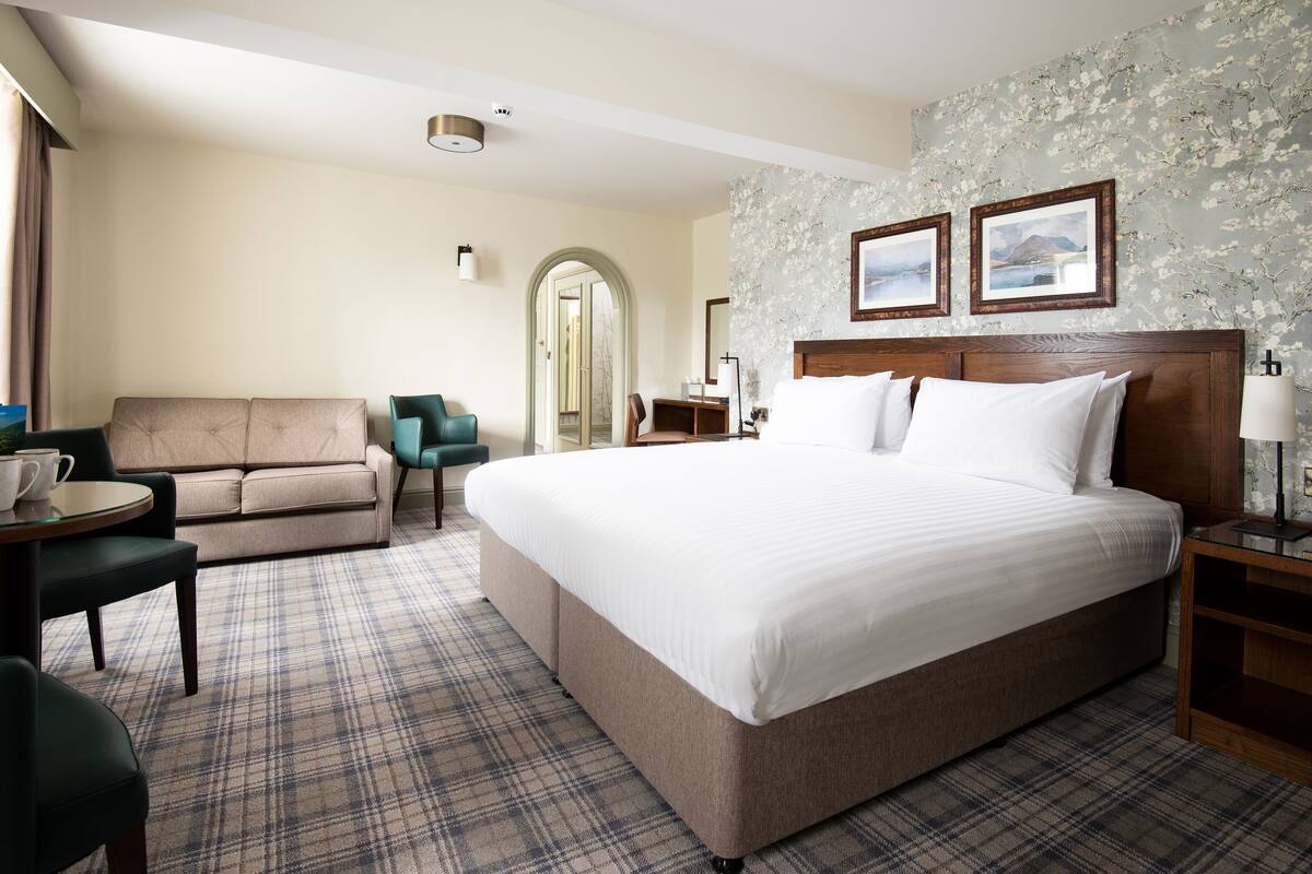 A hotel room with a large bed, a sofa, a small table, and a chair. There is a wooden nightstand with a lamp and a picture frame.