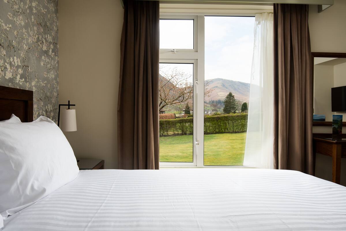 A hotel room with a large bed, a nightstand, a lamp, and a view of a garden and mountains through a sliding door.