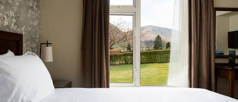 A hotel room with a large bed, a nightstand, a lamp, and a view of a garden and mountains through a sliding door.