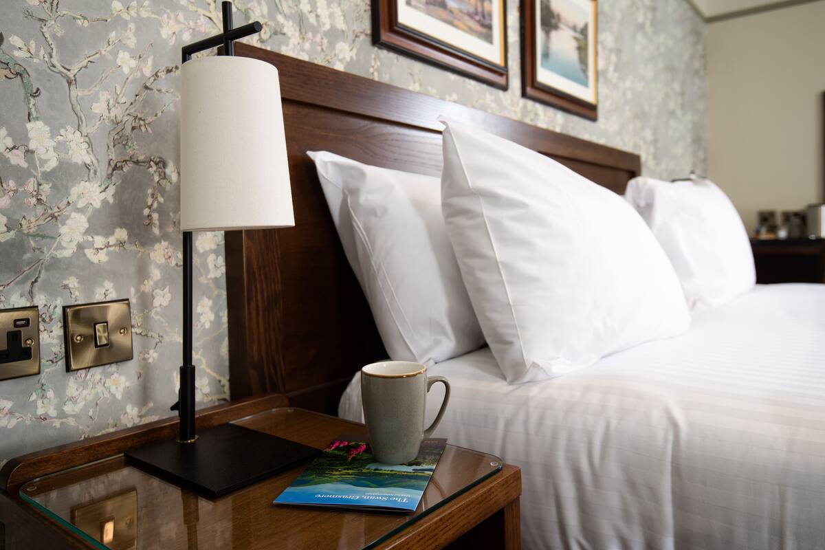 A neatly made bed with white linens, a bedside table with a lamp, a mug, and a book.