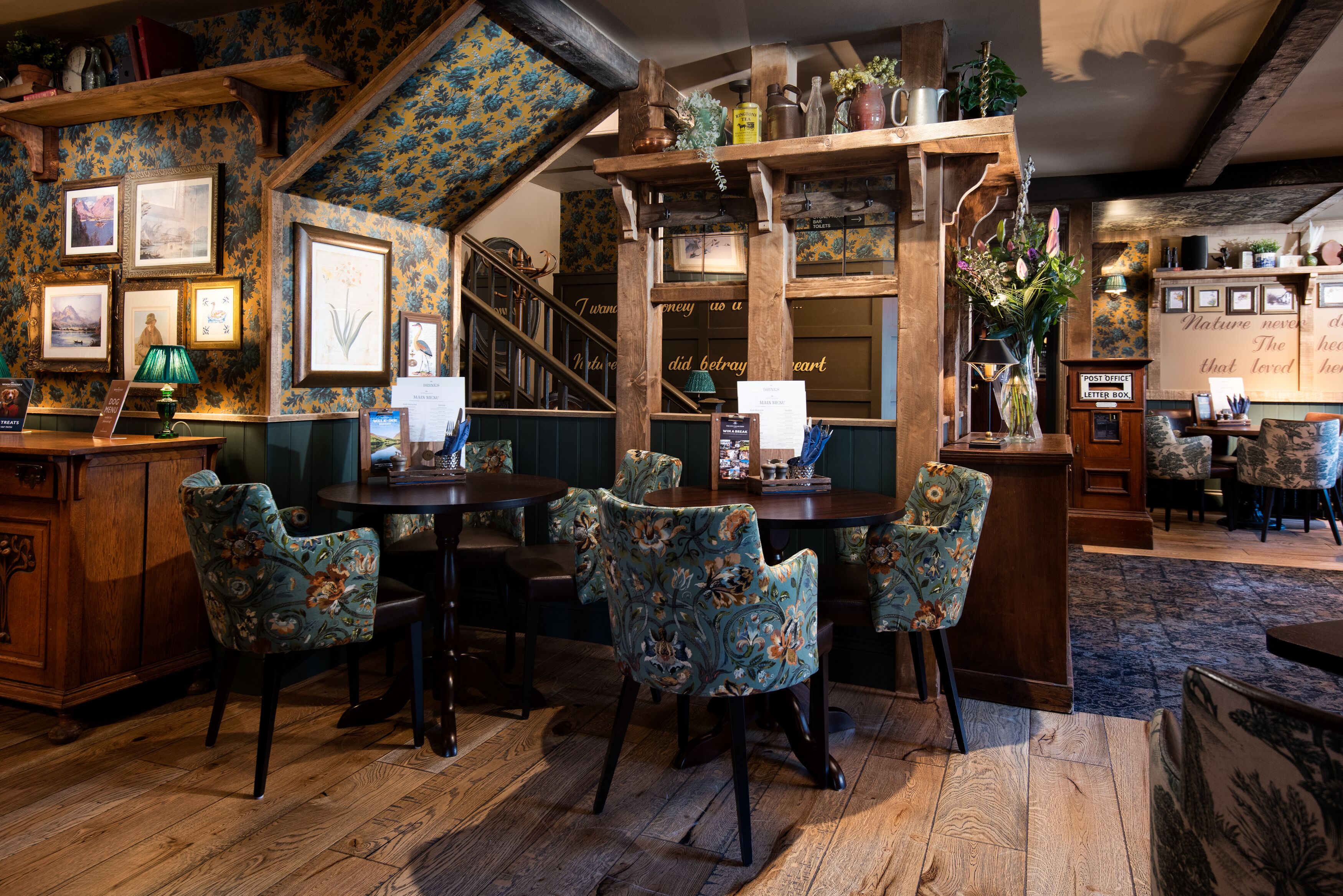 A cozy restaurant with wooden furniture, floral upholstery, and a staircase leading to an upper level.