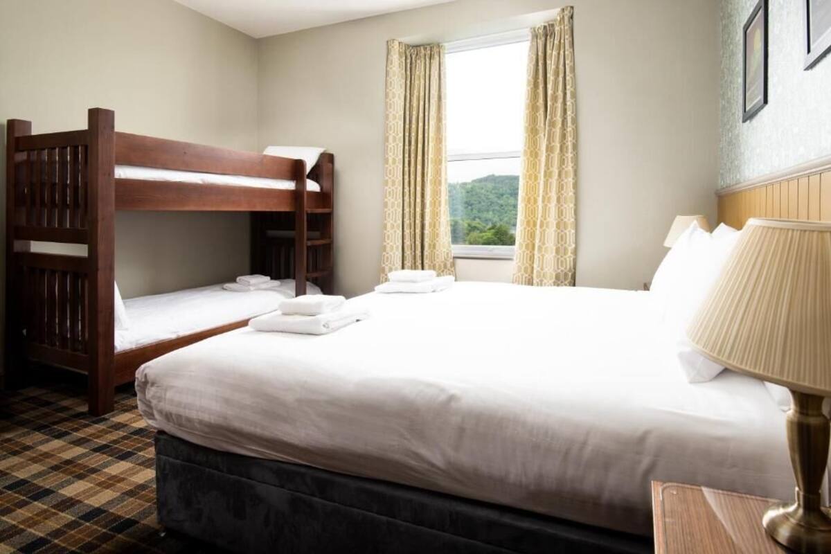 A hotel room with a large bed, a wooden bunk bed, a window with curtains, a bedside lamp, and a checkered floor.