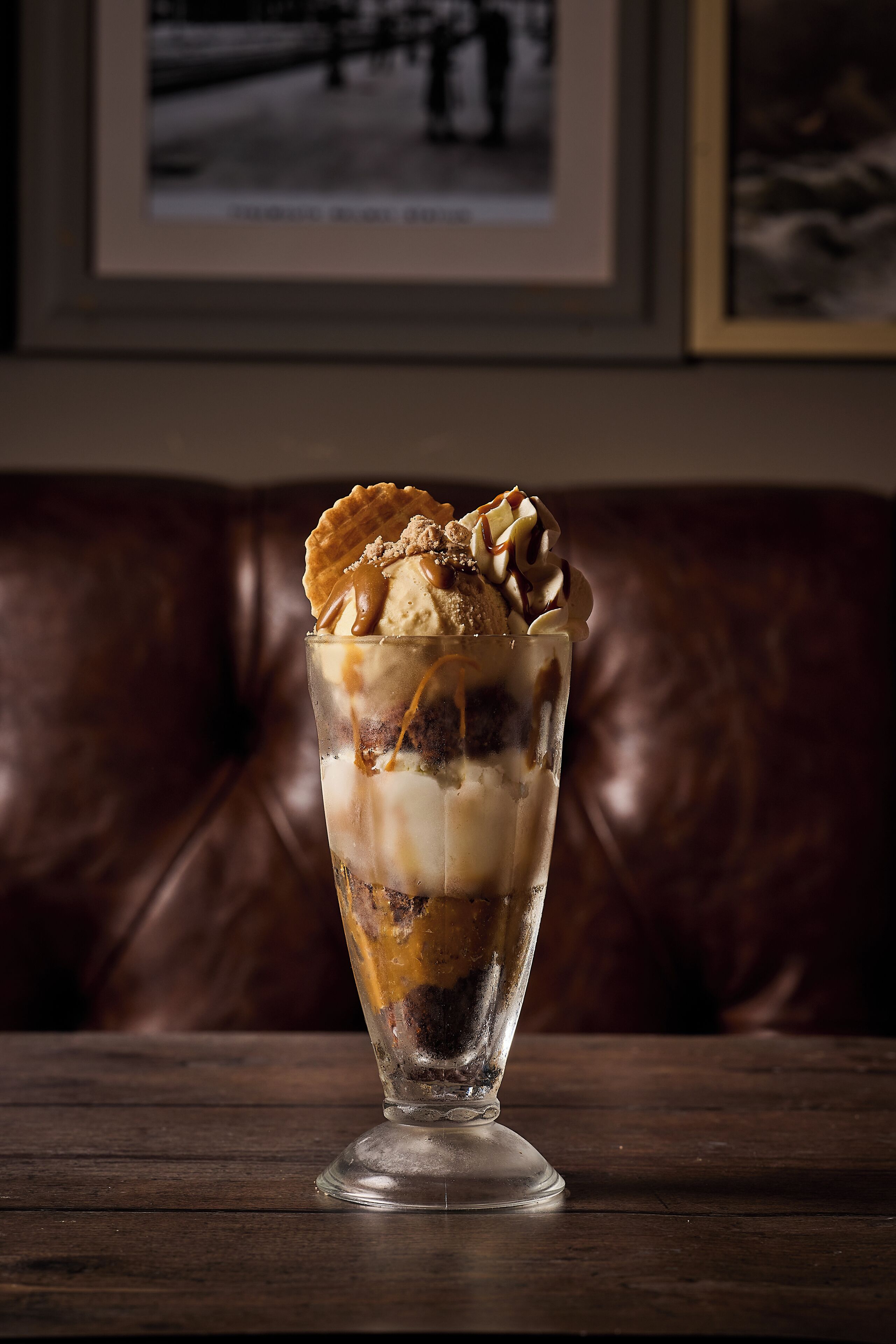 A dessert in a tall glass on a wooden table with a brown leather couch in the background.
