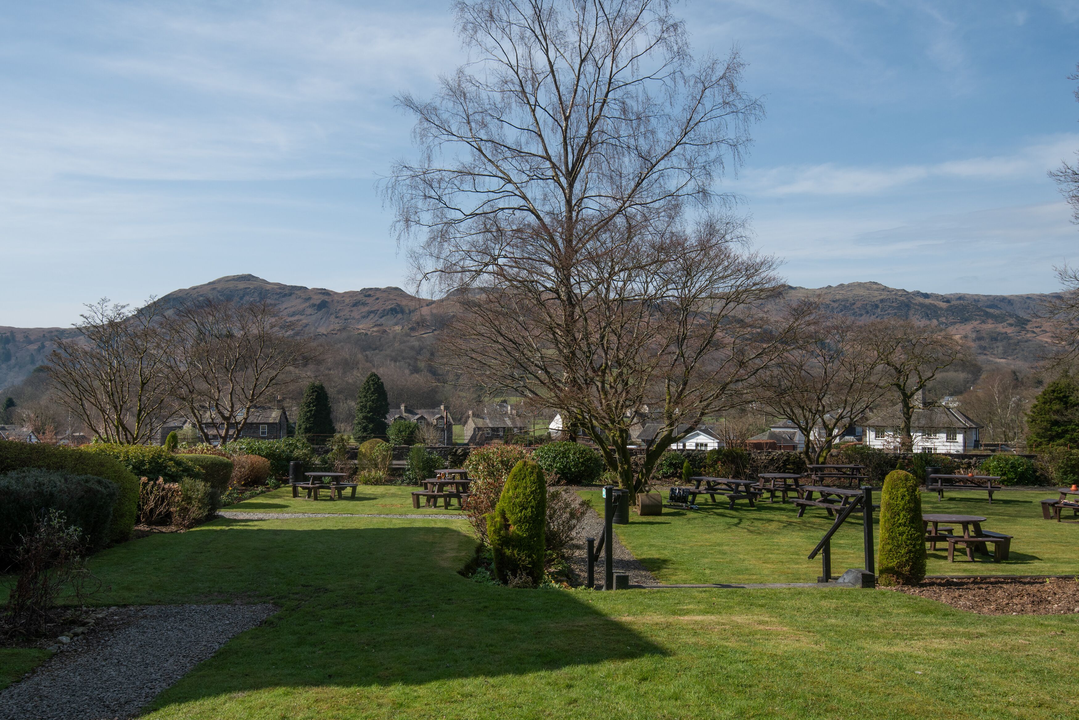 Un parque con mesas de picnic y bancos, rodeado de colinas y casas al fondo.