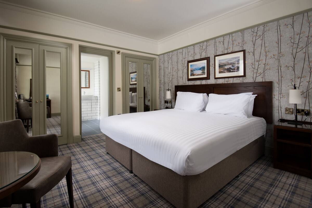 A hotel room with a large bed, a dining table, and a chair. There are two doors leading to other rooms.