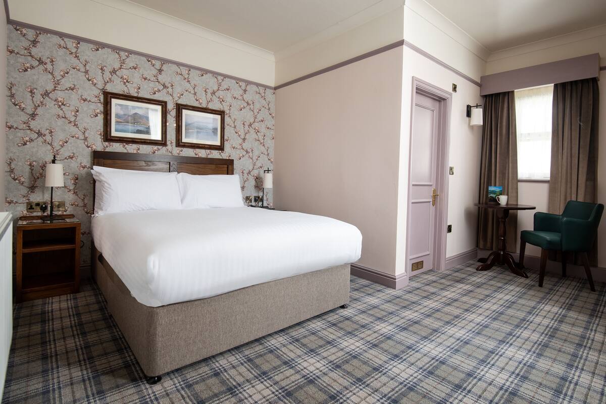 A hotel room with a large bed, a bedside table with a lamp, a chair, a door, and a window with curtains.